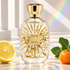 Atelier des Ors Blanc Polychrome on pale stone beside lemon, mandarin, and a fig leaf, with soft rainbow light across a white architectural background.