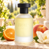 Bois 1920 Classic Paradise frosted bottle beside an orange half, ripe peach, and white magnolia on sunlit stone in a softly blurred garden.