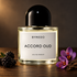 Byredo Accord Oud beside blackberries, purple saffron flowers, and oud wood on a dark wooden surface in warm amber light.
