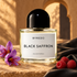 Byredo Black Saffron on sunlit sandstone beside purple saffron crocuses, raspberries, and folded black leather, with an ochre arch behind it.