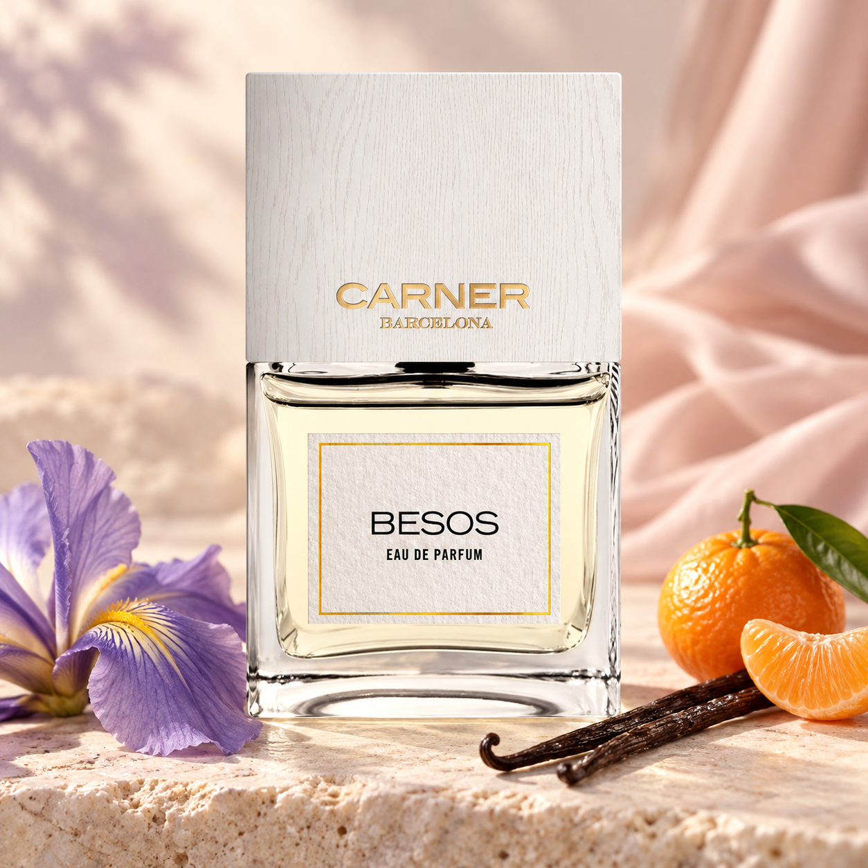 Carner Barcelona Besos beside purple iris, mandarin, and vanilla pods on ivory stone, with blush silk and soft golden light.