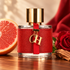 arolina Herrera CH with its red leather wrap and gold charms, beside a grapefruit half, red rose, and cedar wood in warm sunlight.