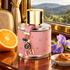 Carolina Herrera CH Grand Tour on a polished table inside a vintage train, beside an orange half, purple iris, and dulce de leche, with green hills outside the window.
