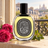 Diptyque Eau Capitale Eau de Parfum bottle beside pink roses and green leaves on a sunlit Parisian stone windowsill.