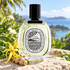Diptyque Eau Mohéli bottle with yellow ylang-ylang flowers and ginger on pale stone overlooking lush tropical greenery and blue sea.