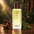 Hermès Agar Ébène beside dark agarwood and green fir branches on a wooden surface, illuminated by warm light against a blurred woodland background.