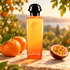 Hermès Eau de Mandarine Ambrée orange bottle beside whole mandarins and a passionfruit half on warm sunlit orchard stone.