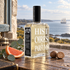 Histoires de Parfums 1828 with eucalyptus, grapefruit, and nutmeg on a weathered wooden table overlooking the coast and a distant sailing ship.