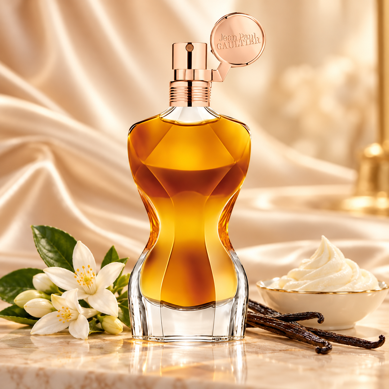 Jean Paul Gaultier Classique Essence de Parfum’s amber faceted bottle beside orange blossoms, vanilla pods, and Chantilly cream against softly lit champagne satin.