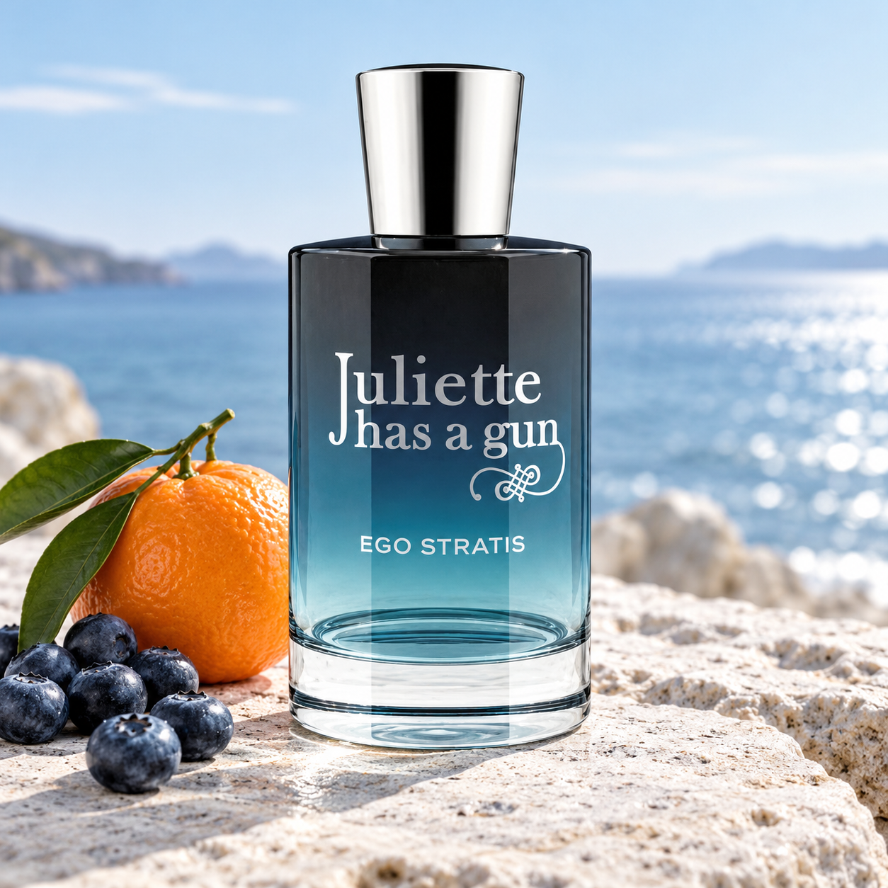Juliette Has A Gun Ego Stratis bottle with a whole tangerine and blueberries on pale stone beside a blue sea.
