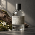 Le Labo AMBROXYDE 17 home fragrance bottle beside weathered wood, green moss, and white jasmine flowers in soft directional light.