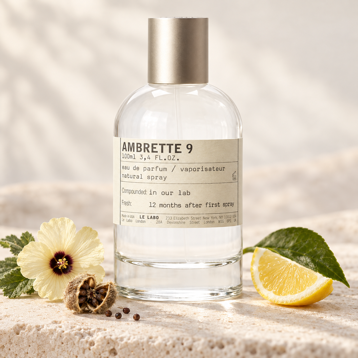 Le Labo AMBRETTE 9 on pale stone beside an ambrette flower, seed pod, and lemon wedge in soft warm light.