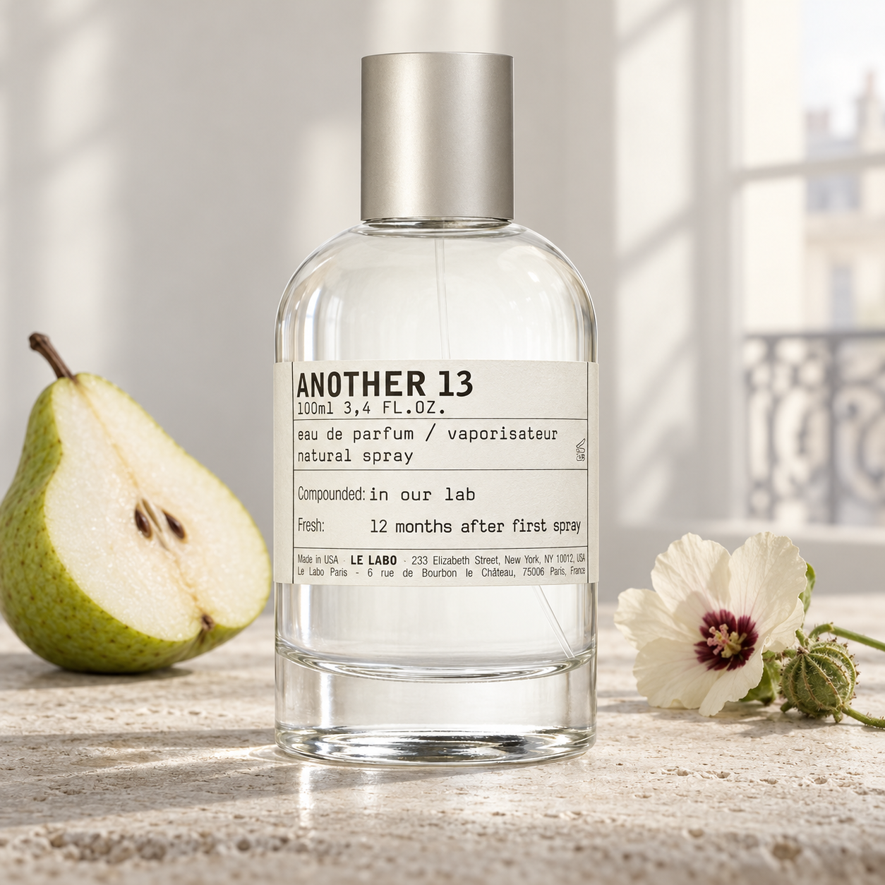 Le Labo Another 13 beside a sliced pear and ambrette flower on pale stone, illuminated by soft window light.