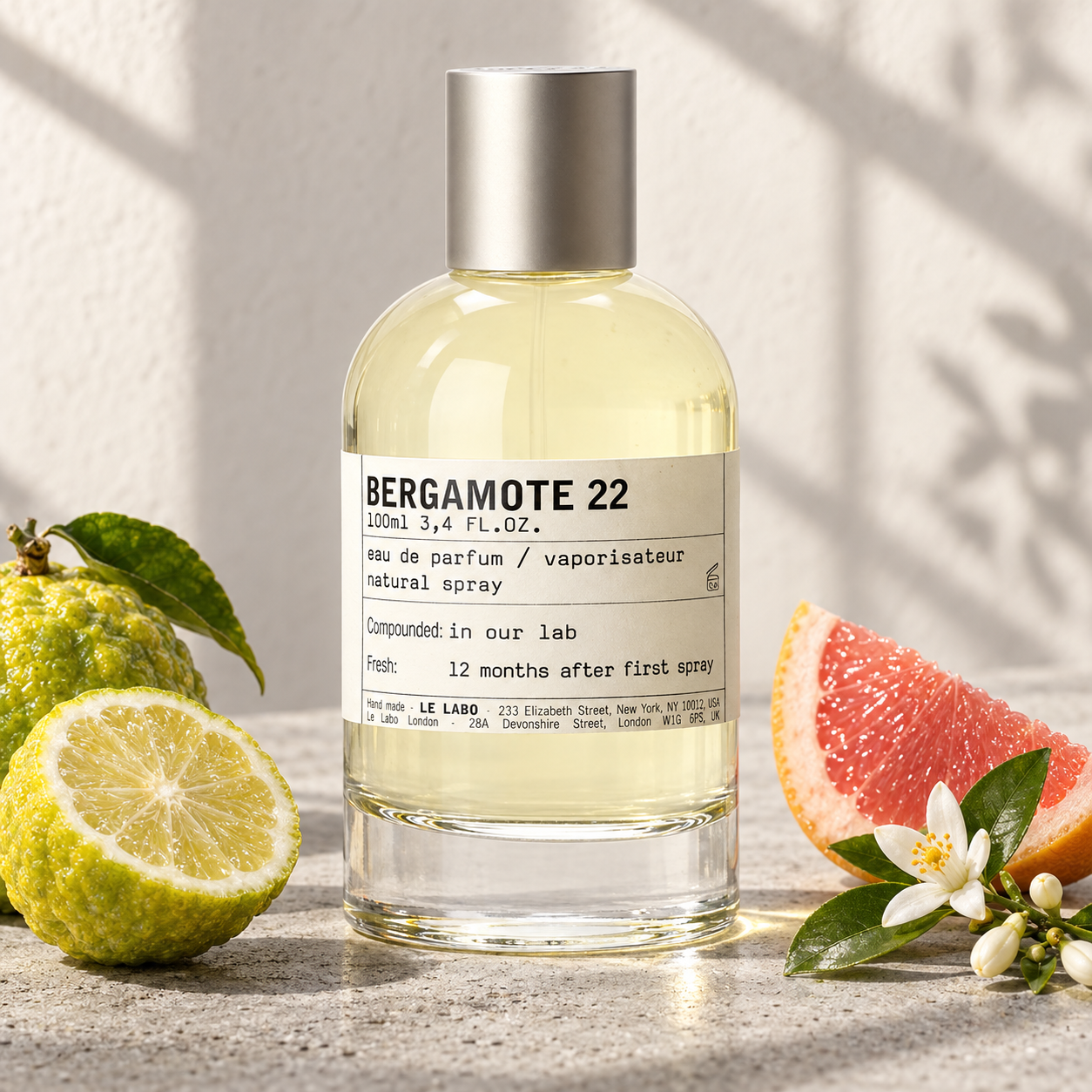 Le Labo Bergamote 22 beside bergamot, pink grapefruit, and orange blossom on pale concrete in soft sunlight.