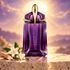 MUGLER Alien’s purple and gold bottle stands on pale stone beside white jasmine and wood, backlit by a golden solar glow.