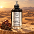Maison Margiela Across Sands perfume beside dates and oud wood on sandstone, with golden desert dunes behind it.