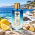 Mancera Intense French Riviera stands on sunlit coastal stone beside lemon, ginger, and sea salt, with turquoise waves and a rocky Mediterranean shoreline behind it