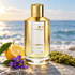 Gold Mancera Amberful bottle beside sliced yuzu, purple violets, and moss on coastal stone, with a softly blurred sea in warm sunlight.