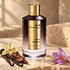 Mancera Aoud Vanille beside vanilla pods, a cream orchid, oud wood, and a saffron crocus on travertine in warm golden light.