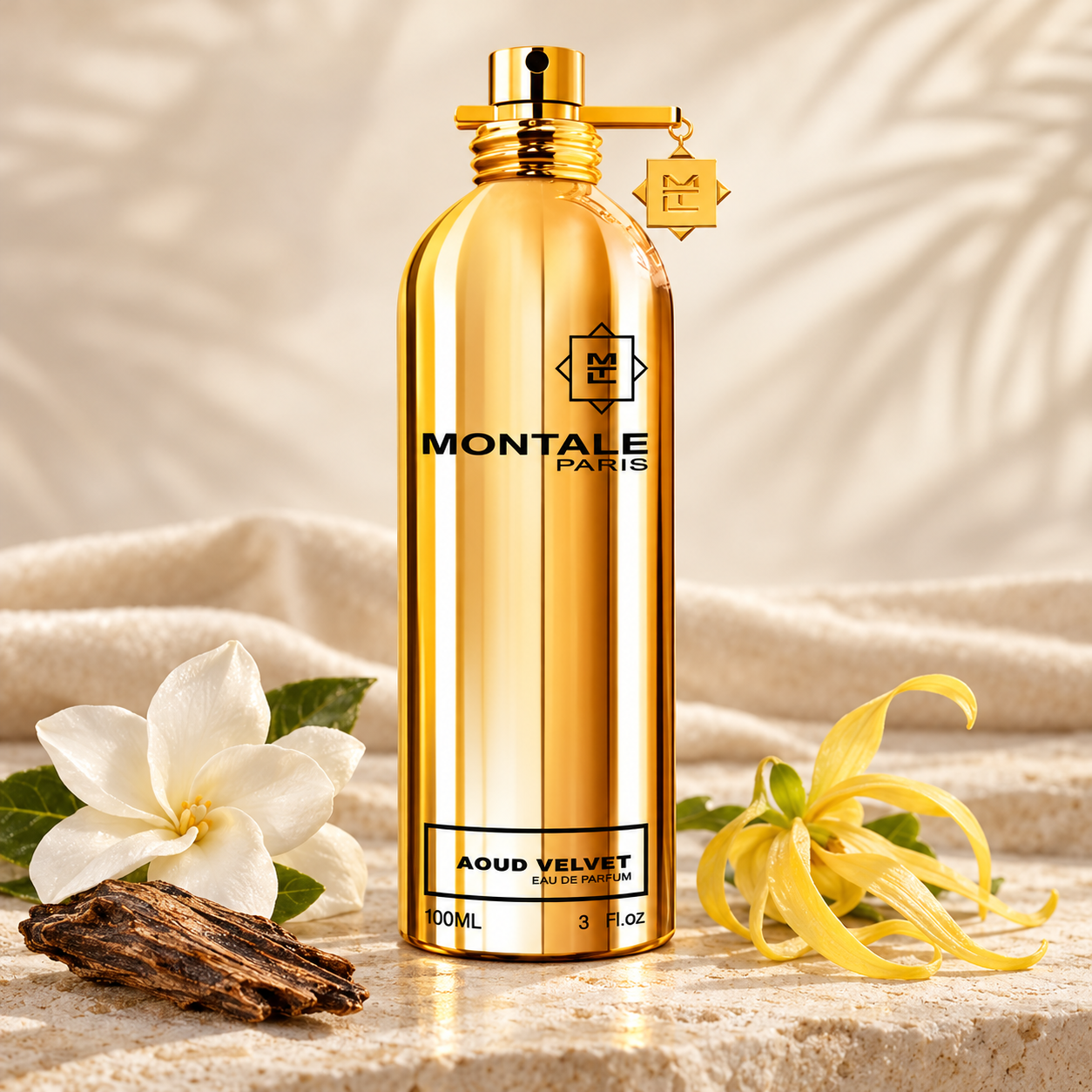 Montale Aoud Velvet’s gold bottle beside white tiare, yellow ylang-ylang, and oud wood on ivory stone with cream velvet behind it.