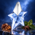 Mugler Angel’s blue star bottle on polished dark blue stone beside praline chocolates, dark chocolate, and green patchouli leaves in cool silver-blue light.