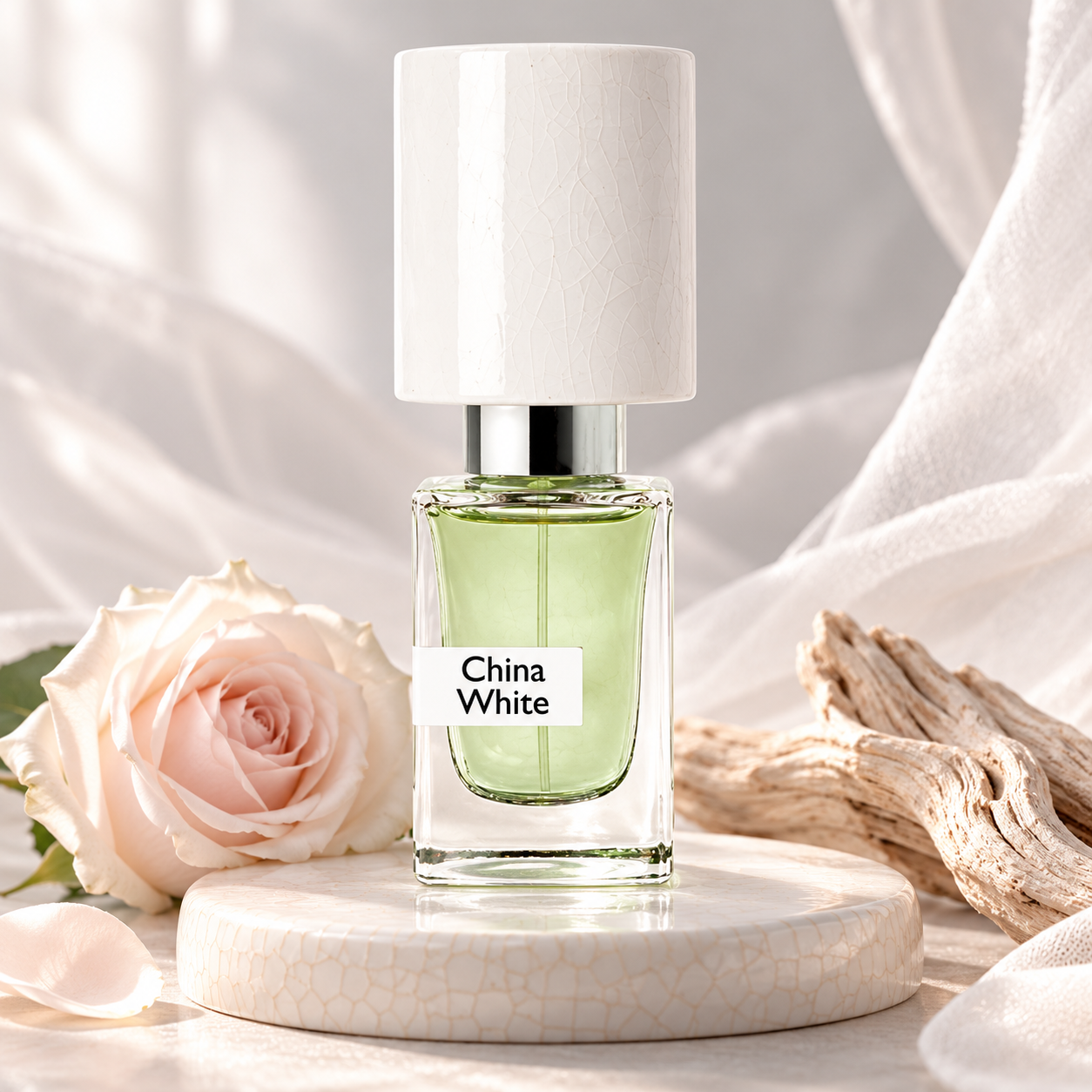 Nasomatto China White on a crackled ivory pedestal beside a pale rose and weathered wood, surrounded by softly draped white fabric.