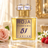 Gold Roja 51 Parfum bottle with a crystal cap beside a pink rose, jasmine, and raspberries on sunlit marble.