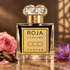 Roja Aoud Parfum’s dark-liquid bottle beside a pink rose, saffron crocus, and oud wood on marble in warm golden light.
