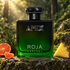 Roja Apex’s green-and-black bottle beside mandarin, pineapple, and cypress on mossy stone in softly lit woodland.