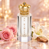 
Royal Crown Al Kimiya’s crystal-clear bottle and jeweled gold crown beside pink roses, a white lily, and sandalwood on softly glowing blush marble.