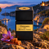 Tiziana Terenzi Chimaera on coastal rock beside saffron flowers, caramel, and pine, overlooking a blue cove with a sailboat and distant flame at dusk.