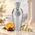 V Canto Agape’s silver bottle on sunlit marble beside orange, chocolate, and white blossoms, with softly blurred classical columns behind it.