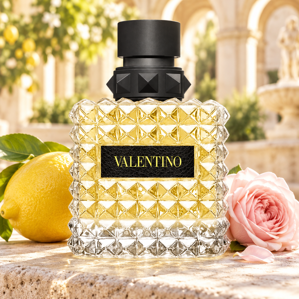 Valentino Donna Born In Roma Yellow Dream bottle with a whole lemon and pale pink rose in a sunny Roman courtyard.