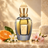 Xerjoff Amber Gold bottle on pale stone beside sliced tangerine, white orange blossom, and pink almond blossoms in golden light.