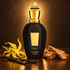 Black and gold Xerjoff Amber Star bottle beside yellow ylang-ylang, vanilla pods, and sandalwood on polished wood in warm golden light.