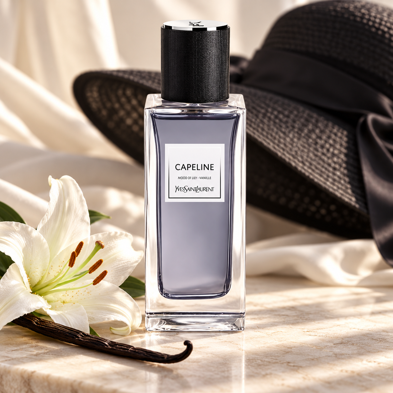 Yves Saint Laurent Capeline with a white lily and vanilla pod on ivory stone, backed by draped fabric and a black wide-brimmed hat.