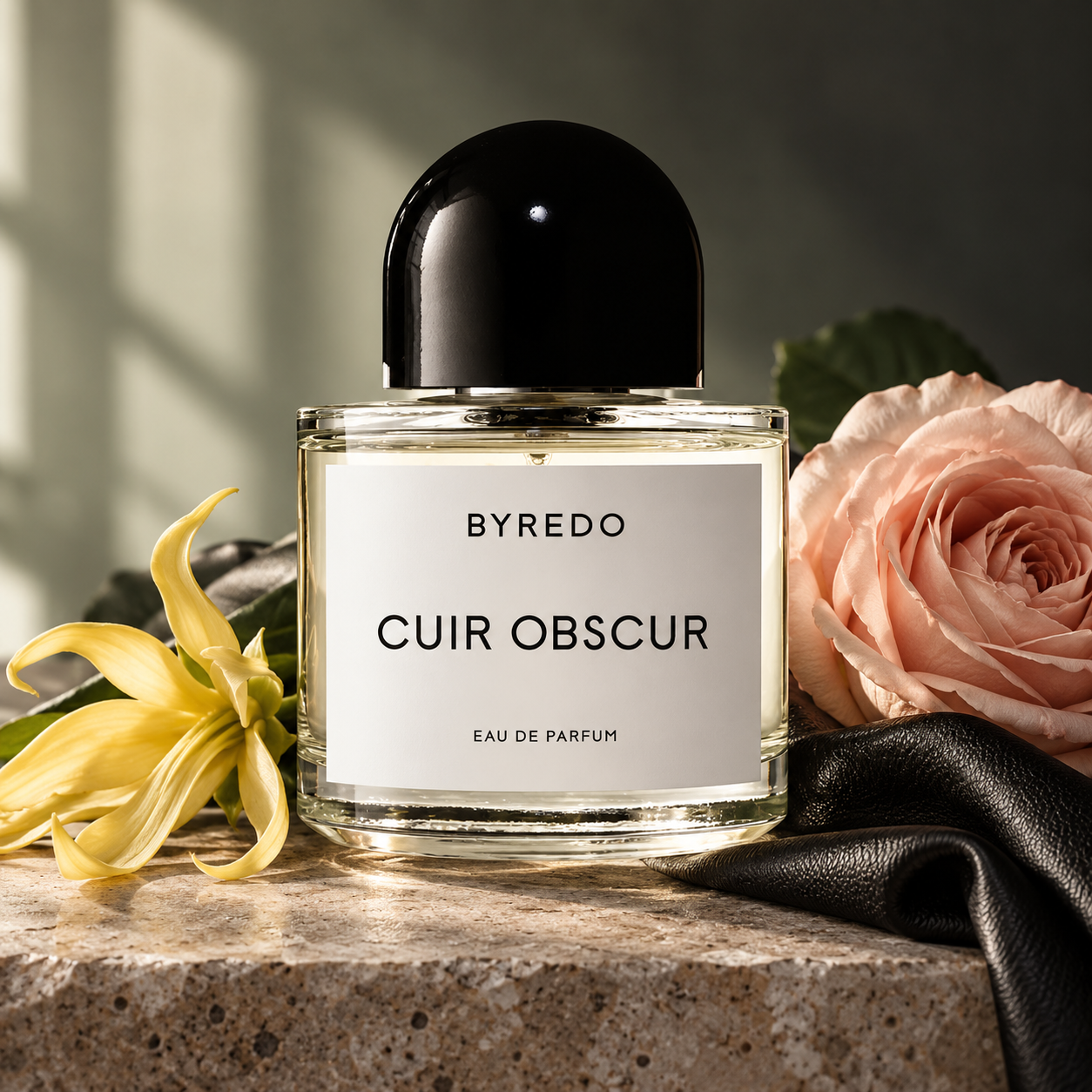 Byredo Cuir Obscur bottle with a black cap beside a blush rose, yellow ylang-ylang flower and black leather on taupe stone.