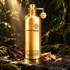 Montale Dark Aoud gold bottle beside oud wood and sandalwood shavings on dark stone in softly lit forest surroundings.
